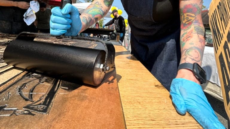 A person's hands and arms with colorful tattoos are rolling black ink onto a woodblock print